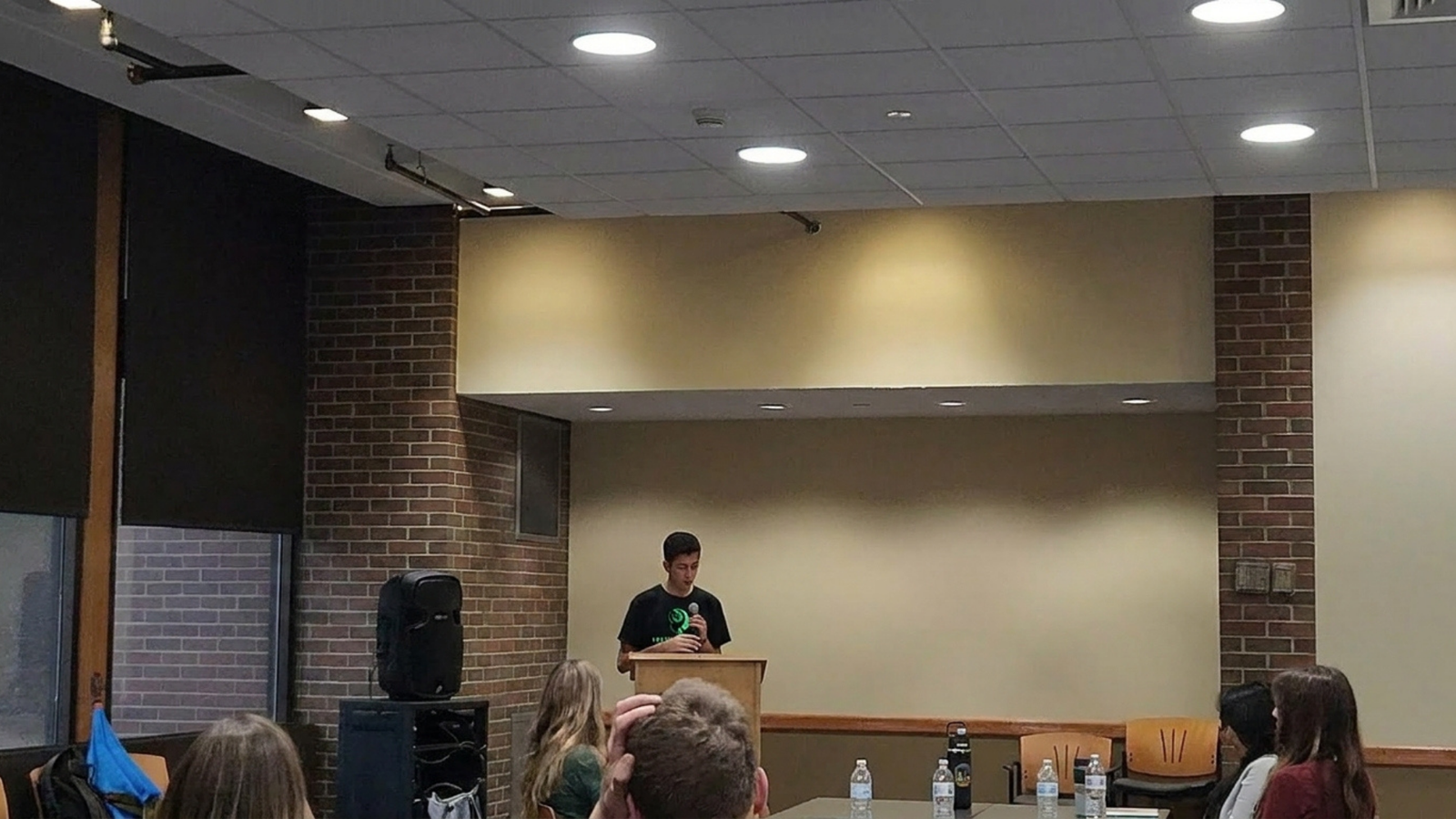 Aditya Herekar speaking at a podium to a seated audience in a college meeting room